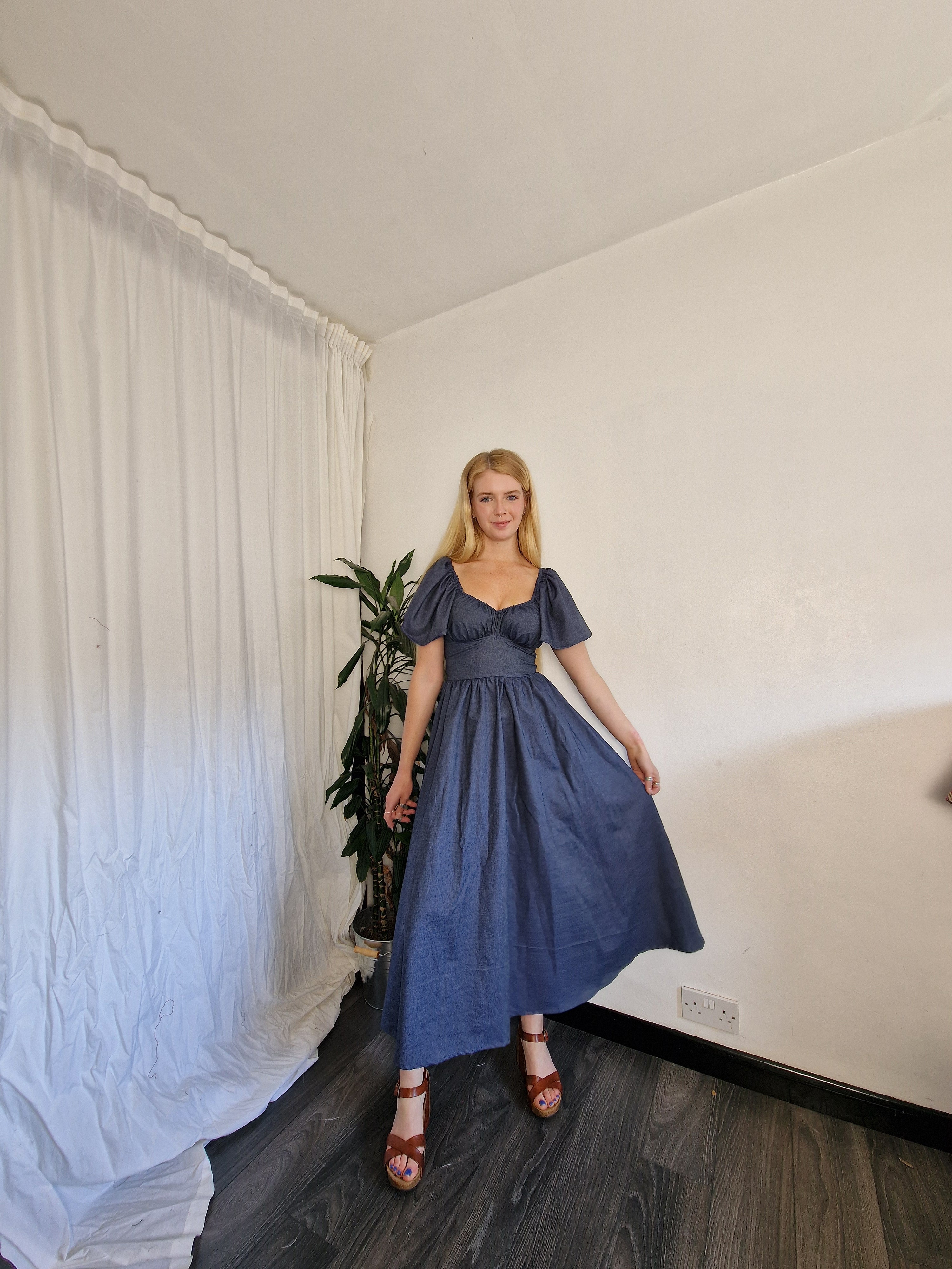 Woman in a blue dress standing in a room with a white wall and dark floor.