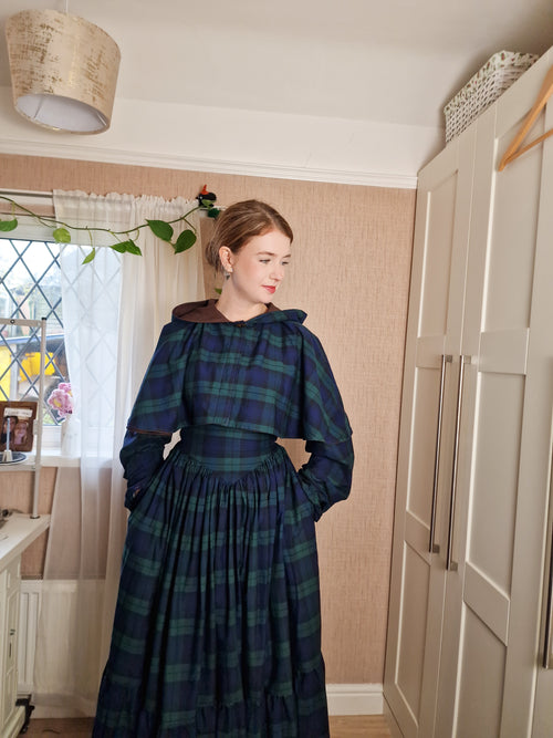 Woman wearing a dark blue plaid dress standing in a room with a window and wardrobe.
